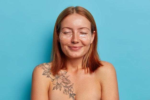 Mujeres, cuidado facial y concepto de belleza. Alegre mujer pelirroja sonriente se para con los ojos cerrados, se aplica parches de hidrogel, tiene una piel suave y limpia, un cuerpo bien cuidado, posa desnuda contra una pared azul