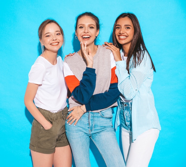 Mujeres despreocupadas atractivas que presentan cerca de la pared azul en estudio. Modelos positivos divirtiéndose y abrazándose