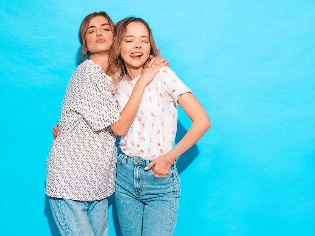 Mujeres despreocupadas sexy posando junto a la pared azul. Modelos positivos divirtiéndose y mostrando lengua