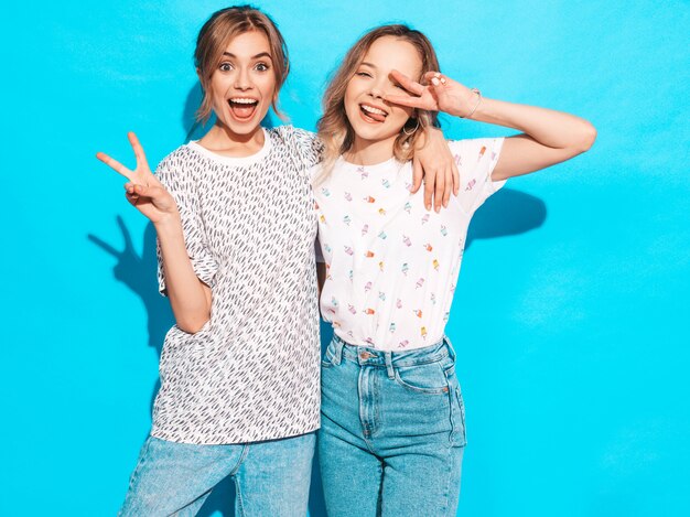 Mujeres despreocupadas sexy posando junto a la pared azul. Modelos positivos que se divierten. Muestran signo de la paz y lengua