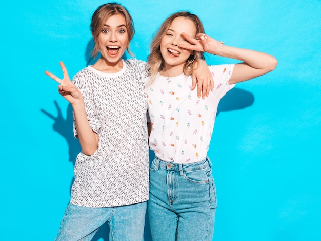 Mujeres despreocupadas sexy posando junto a la pared azul. Modelos positivos que se divierten. Muestran signo de la paz y lengua