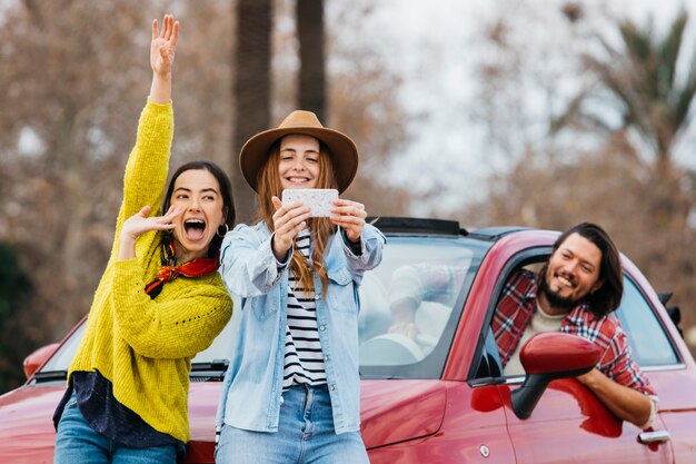 Mujeres divirtiéndose y tomando autofotos en un teléfono inteligente cerca del hombre que se inclina hacia fuera del auto
