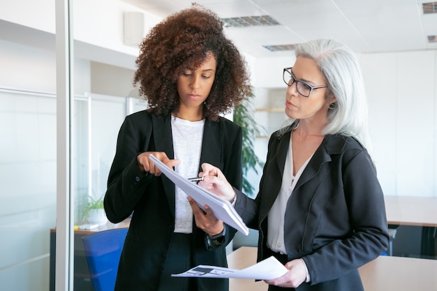 Mujeres empresarias atractivas concentradas que comparan datos analíticos. Profesionales femeninas seguras de éxito leyendo documentos o informes en la sala de reuniones. Concepto de trabajo en equipo, negocios y gestión