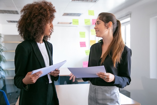 Mujeres empresarias bastante jóvenes discutiendo el plan del proyecto y sonriendo. Dos hermosas colegas sosteniendo documentos y hablando en la sala de conferencias. Concepto de trabajo en equipo, negocios y gestión