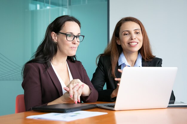 Mujeres empresarias que tienen video charla con socios, sentado en la computadora portátil abierta, mirando la pantalla y sonriendo