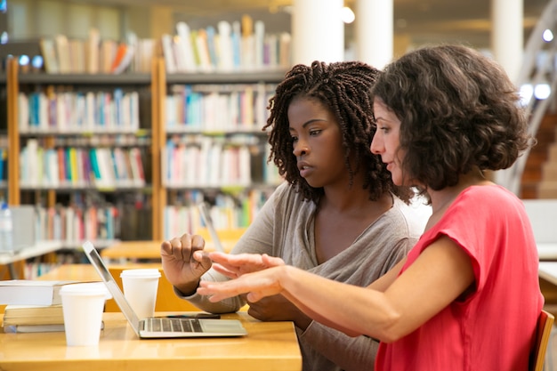 Mujeres enfocadas hablando y apuntando a la computadora portátil en la biblioteca