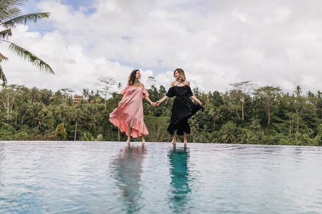 Mujeres espectaculares jugando con sus vestidos mientras posan cerca del lago. Tiro de cuerpo entero al aire libre de señoras de la mano en la naturaleza.