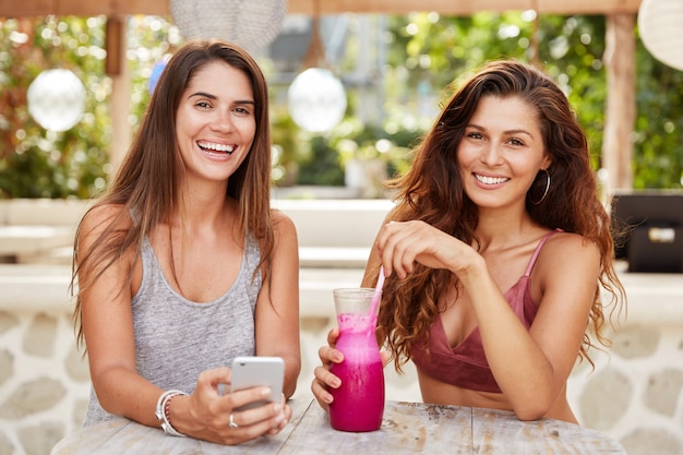 Mujeres felices con miradas felices, divertirse juntas, leer comentarios en blogs en teléfonos inteligentes, tomar bebidas frescas, sentarse en la cafetería al aire libre.