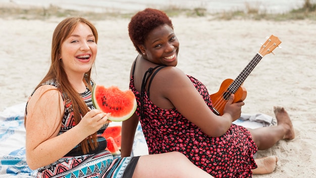 Foto gratuita mujeres felices posando en la playa con sandía y guitarra