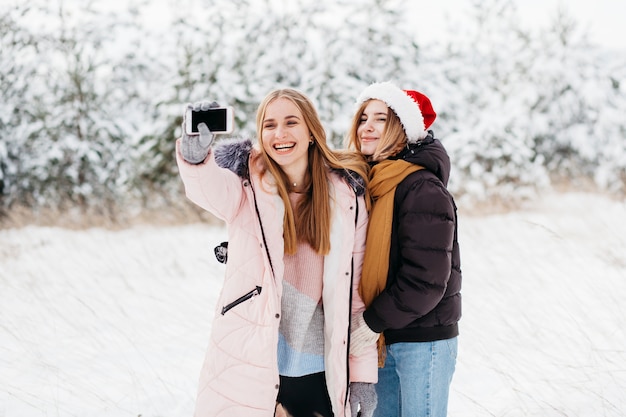 Mujeres felices en Santa sombrero tomando selfie en bosque del invierno