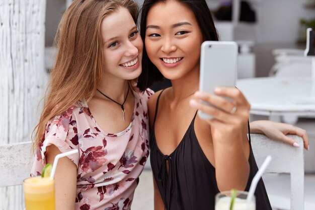 Mujeres felices con sonrisas agradables toman fotos en el teléfono celular, pasan tiempo libre juntas en la cafetería y beben cócteles frescos, disfrutan de las vacaciones en el lugar del resort.