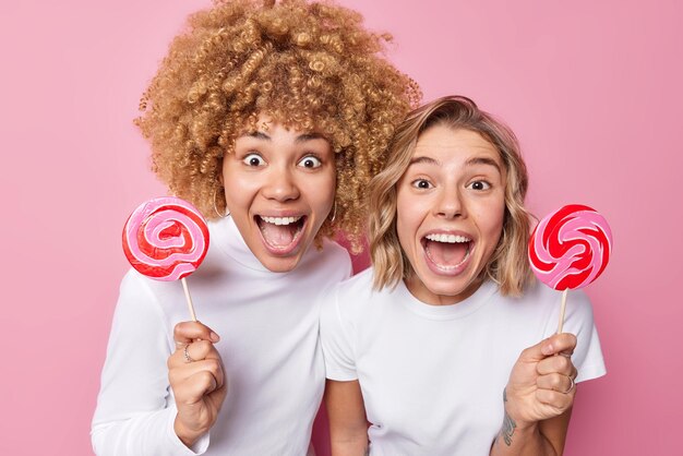 Las mujeres felices y sorprendidas miran con gran asombro a la cámara, mantienen la boca abierta, se sienten emocionadas, sostienen los dulces de caramelo, tontas, vestidas casualmente, aisladas sobre fondo rosa. Wow, qué piruleta sabrosa.