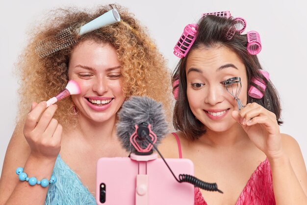 Las mujeres hacen maquillaje aplicando polvo facial usar pestañas rizador grabar video blog compartir en las redes sociales pararse frente al teléfono inteligente hacer peinado aislado en blanco