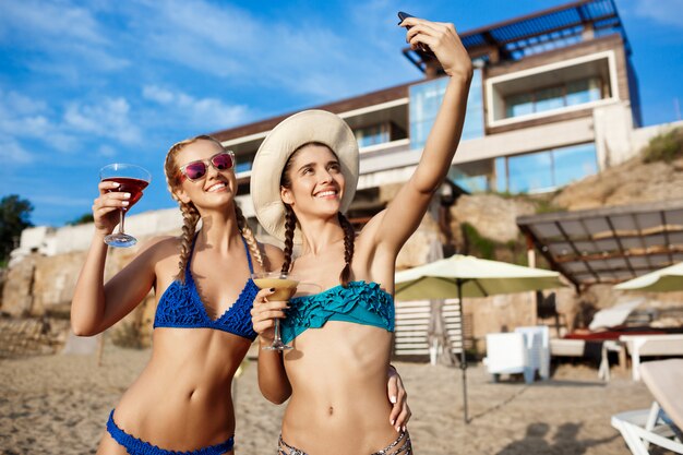 Mujeres hermosas jóvenes en traje de baño sonriendo, haciendo selfie en la playa