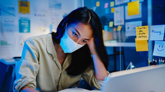 Las mujeres independientes de Asia usan mascarilla usando una computadora portátil en el trabajo duro en la nueva oficina normal. Trabajar desde casa sobrecarga nocturna, autoaislamiento, distanciamiento social, cuarentena para la prevención del virus corona.