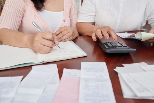 Mujeres irreconocibles sentadas a la mesa con recibos, contando con calculadora y escribiendo en diario