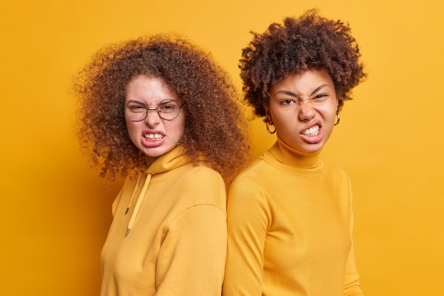 Las mujeres irritadas tienen malentendidos y se pelean, caras sonrientes, apretar los dientes, mirar con enojo, ponerse de espaldas, sentirse molestas, aisladas sobre la pared amarilla. Relación de desacuerdo