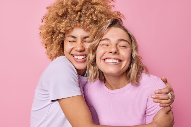 Las mujeres jóvenes alegres se abrazan con amor, se sienten muy contentas, la sonrisa muestra ampliamente que los dientes blancos se preocupan unas por otras, vestidas con camisetas informales aisladas sobre un fondo rosa Concepto de amistad