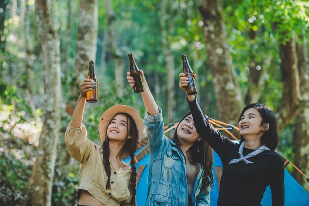 Mujeres jóvenes y amigas viajeras relajándose en sillas de campamento en la tienda Están animando y bebiendo cerveza durante el campamento hablando con diversión y felices juntos