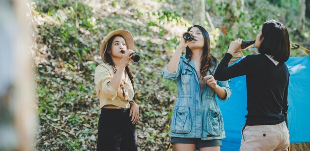 Mujeres jóvenes y amigas viajeras relajándose en sillas de campamento en la tienda Están animando y bebiendo cerveza durante el campamento hablando con diversión y felices juntos