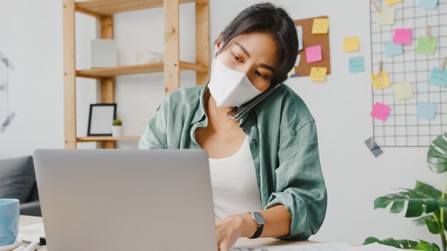 Las mujeres jóvenes de Asia usan mascarilla médica hablando por teléfono, empresario ocupado que trabaja a distancia en la sala de estar.