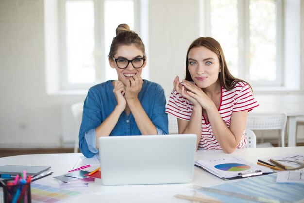 Mujeres jóvenes emocionadas mirando felizmente en la cámara trabajando juntas con una computadora portátil mientras pasan tiempo en la oficina moderna