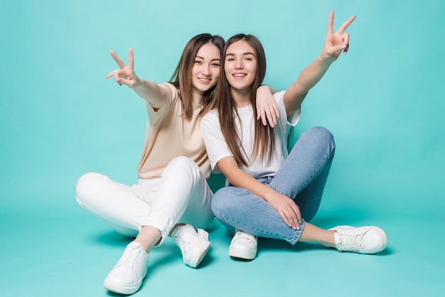 Mujeres jóvenes emocionadas con paz posando en el piso aislado en la pared turquesa.