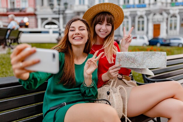 Mujeres jóvenes con estilo que viajan juntas vestidas con vestidos y accesorios de moda de primavera que se divierten tomando fotos en la cámara del teléfono sosteniendo el mapa