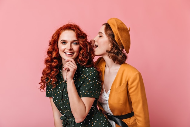 Mujeres jóvenes eufóricas hablando sobre fondo rosa. Foto de estudio de dos amigos en traje vintage.