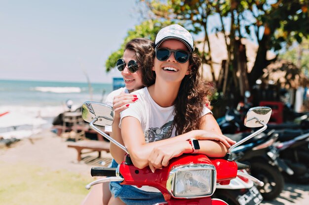 Mujeres jóvenes felices explorando la isla en motocicleta, con sombreros de verano, usando tableta y comprando música en línea contra el fondo de la ciudad, isla exótica, viaje, vacaciones de verano