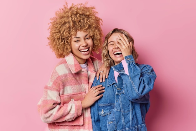 Las mujeres jóvenes felices y llenas de alegría se ríen alegremente al escuchar algo muy divertido vestidas con ropa de moda se paran cerca unas de otras aisladas sobre un fondo rosa Concepto de amistad y emociones