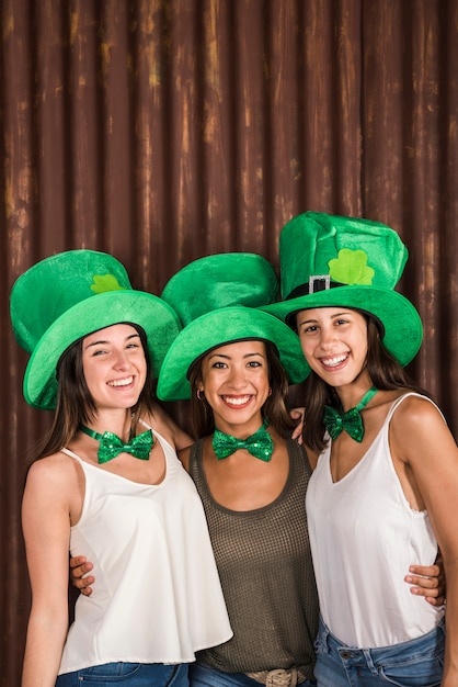 Mujeres jovenes felices en los sombreros de San Patricio que abrazan cerca de la pared