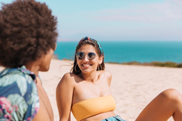 Mujeres jóvenes en gafas de sol mirando chico y sonriendo en la playa