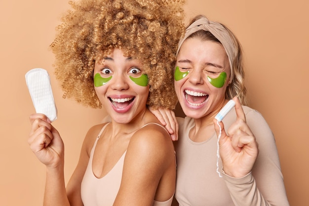 Las mujeres jóvenes positivas sonríen alegremente, sostenga de cerca la toalla sanitaria y el tampón elija el mejor producto para la menstruación aplique parches de hidrogel verde debajo de los ojos para el tratamiento de la piel Concepto de belleza