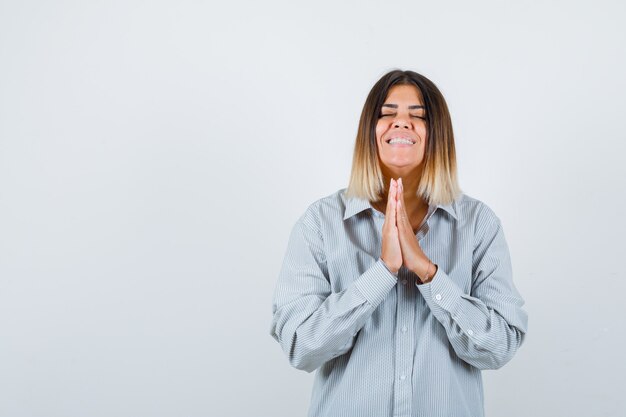 Las mujeres jóvenes presionan las manos juntas para orar con una camisa de gran tamaño y lucir feliz, vista frontal.