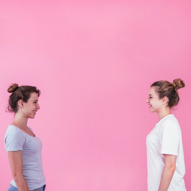 Mujeres jóvenes sonrientes de pie cara a cara mirando el uno al otro sobre fondo rosa