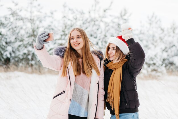 Mujeres lindas en Santa sombrero tomando selfie en bosque de invierno