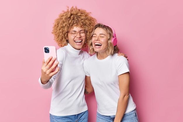 Las mujeres llenas de alegría posan para selfie ríen alegremente divertidas sostienen el teléfono inteligente delante vestidas con ropa informal usan auriculares estéreo aislados sobre fondo rosa Dos amigos se hacen fotos a sí mismos