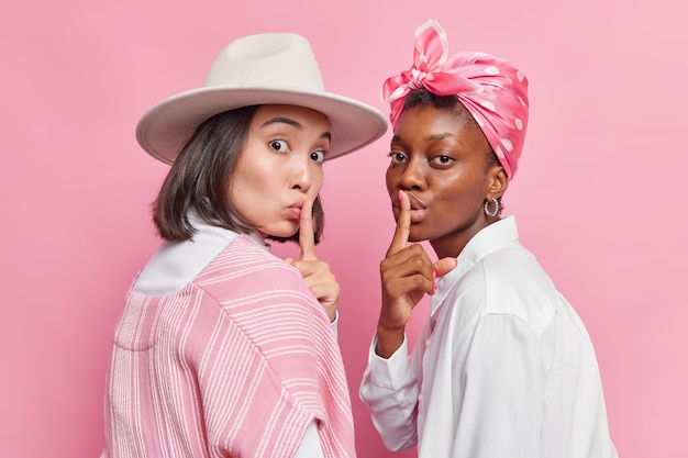 las mujeres miran en secreto a la cámara esconden algo piden que se callen hagan un gesto de silencio usar ropa elegante posar una al lado de la otra aislado en rosa