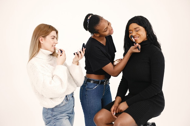 Mujeres mixtas maquillando a su novia. Amigos multirraciales posando aislado sobre fondo de pared blanca.