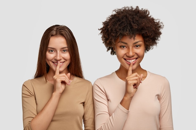 mujeres multiétnicas divertidas positivas muestran el signo de silencio, piden que alguien se calle, difunden rumores, tienen expresiones felices