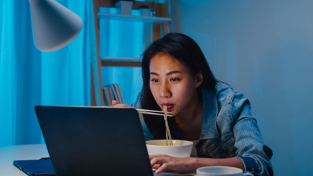 Mujeres de negocios inteligentes independientes de Asia que comen fideos instantáneos mientras trabajan en la computadora portátil en la sala de estar en casa por la noche. La muchacha asiática joven feliz que se sienta en el escritorio trabaja horas extras, disfruta del tiempo de la relajación.