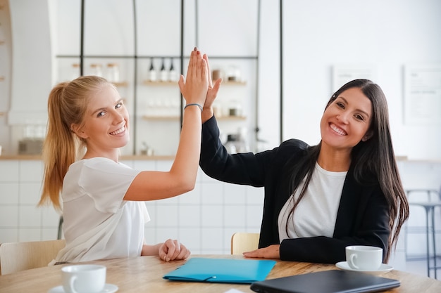 Foto gratuita mujeres de negocios jóvenes felices dando cinco y celebrando el éxito, sentados a la mesa con documentos y tazas de café, mirando a la cámara