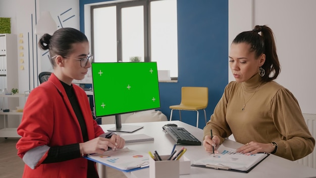 Mujeres de negocios usando computadora con pantalla verde en el escritorio. Equipo de personas hablando mientras trabajan con un fondo simulado y una plantilla aislada en la pantalla de la computadora. Espacio de copia de clave de croma