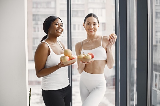 Mujeres negras y caucásicas de pie en un estudio con grandes ventanas y sosteniendo manzanas