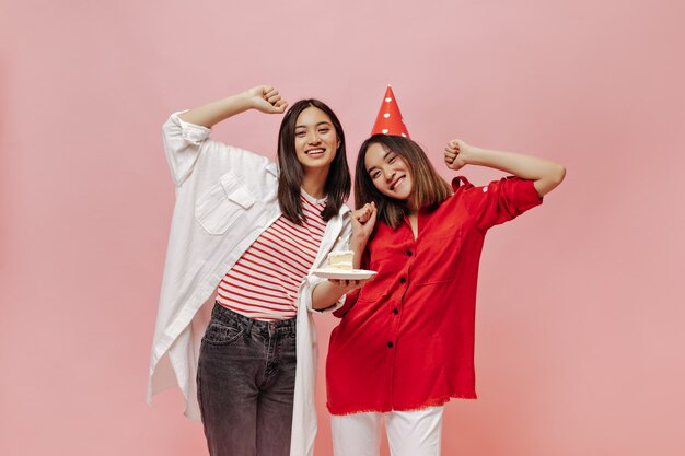 Las mujeres de pelo corto celebran el cumpleaños en la pared rosa aislada. Encantadora chica en camiseta a rayas y camisa de gran tamaño sostiene b-day cake