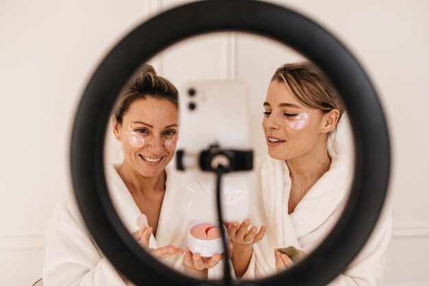 Las mujeres de piel bastante clara usan parches antiarrugas y toman fotos a través de una lámpara de anillo en un trípode. Procedimiento de belleza, concepto de ritual matutino.