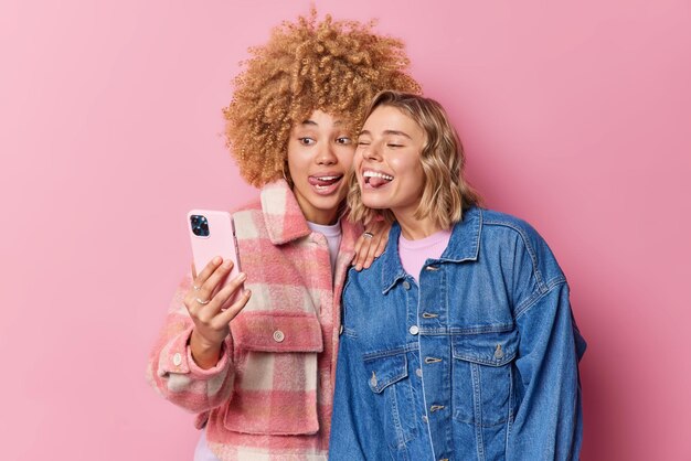Las mujeres positivas y alegres hacen una mueca ante la cámara del teléfono inteligente sacan la lengua tontamente toman selfie vestidas con ropa de moda aisladas sobre fondo rosa hacen una foto de sí mismas