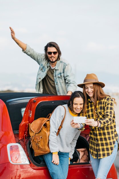 Mujeres positivas que toman autofotos en un teléfono inteligente cerca del maletero del coche y el hombre que se inclina hacia fuera del auto