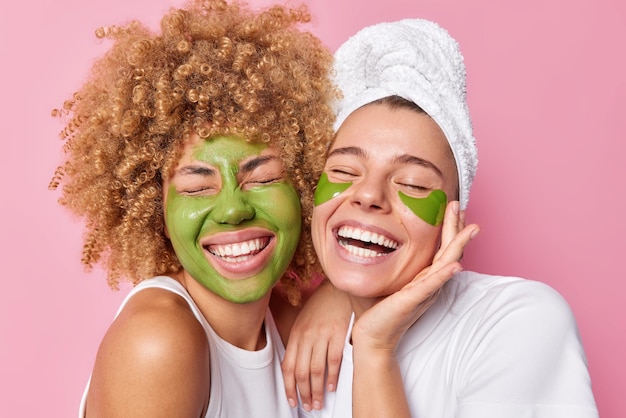Las mujeres positivas sienten una sonrisa alegre, mantienen los ojos cerrados, aplican una máscara nutritiva verde y parches vestidos con camisetas blancas se someten a procedimientos de belleza aislados sobre fondo rosa Tratamiento facial